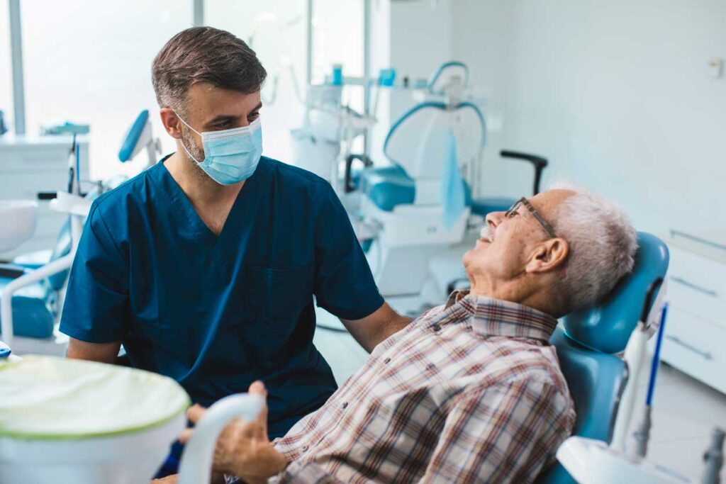 A dentist in blue scrubs and a face mask is sitting beside an elderly man in a dental chair. They are having a friendly conversation in a brightly lit dental office.